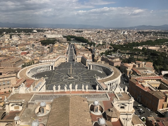 An expansive view of a grand cityscape featuring St. Peter's Square in the Vatican City, surrounded by historical buildings with columns and statues. Roads and pathways extend outwards, and a central obelisk is prominent within the square. Behind the square, the city's architecture continues with a mix of historical and modern buildings, with verdant areas visible in the distance under a partly cloudy sky.