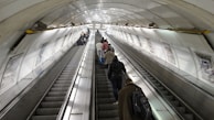 A long underground escalator tunnel with people using both sides. The walls are lined with advertisements and the lighting is bright, reflecting off the metallic surfaces. The escalator leads upwards, and the ceiling is curved with a series of lights along the top.
