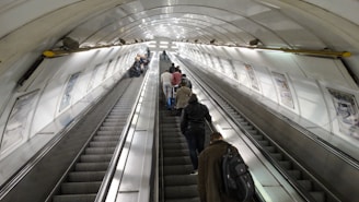 A long underground escalator tunnel with people using both sides. The walls are lined with advertisements and the lighting is bright, reflecting off the metallic surfaces. The escalator leads upwards, and the ceiling is curved with a series of lights along the top.
