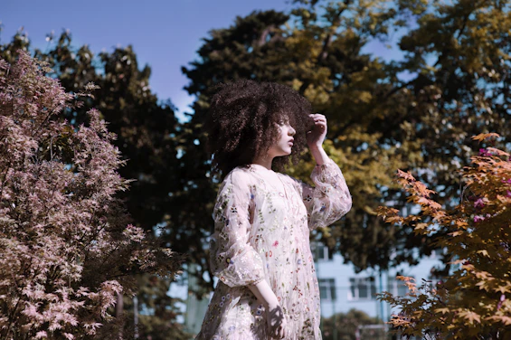 Model wearing a flowing floral dress standing in a sunlit garden with soft shadows.