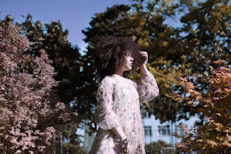 A model wearing a flowing neutral-toned dress standing in a sunlit garden with soft pink roses.