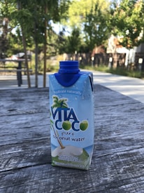 A carton of Vita Coco pure coconut water is placed on a weathered wooden surface. The carton is adorned with images of palm trees and coconuts and features a blue screw cap. In the background, there's a serene park setting with trees, benches, and a pathway.