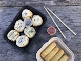 A set of steamed buns with black sesame seeds in a black rectangular container and spring rolls in a beige container are laid out on a wooden table. Two small containers of sauces, likely soy and chili, are included next to a pair of wooden chopsticks.