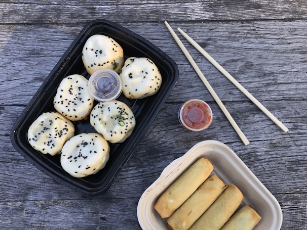 A set of steamed buns with black sesame seeds in a black rectangular container and spring rolls in a beige container are laid out on a wooden table. Two small containers of sauces, likely soy and chili, are included next to a pair of wooden chopsticks.
