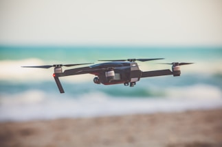 A black quadcopter drone is flying near a beach, with a blurred view of the ocean in the background. It is an outdoor setting with a sunny atmosphere.