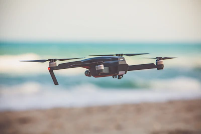 A sturdy quadcopter lifting off from a sandy beach with ocean waves behind.