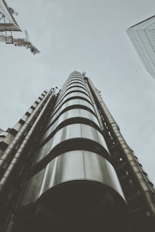 A futuristic building with a focus on its metallic, cylindrical structure rising toward the sky. The architecture is industrial and modern, featuring exposed pipes and a sleek design. Cranes are visible in the sky, indicating ongoing construction or development nearby.
