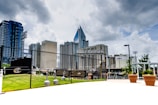 A modern urban skyline dominates the scene with tall skyscrapers and a prominent glass building in the center. In the foreground, a chain-link fence and a sign reading 'BB&T BallPark' indicate the location of a sports field. The atmosphere is enhanced by dramatic cloud formations in the sky, contrasting with the bright green grass and the urban setting.