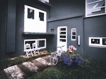 A modern exterior of a building with dark walls featuring white-framed doors and windows. On the left, two white chairs sit on a small veranda. A bicycle leans against the wall near the entrance door surrounded by vibrant flower pots. The ground is a mix of grass and paved stepping stones.