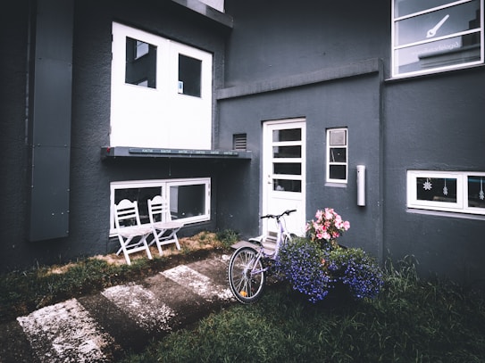 A modern exterior of a building with dark walls featuring white-framed doors and windows. On the left, two white chairs sit on a small veranda. A bicycle leans against the wall near the entrance door surrounded by vibrant flower pots. The ground is a mix of grass and paved stepping stones.