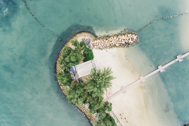 An aerial view of a small peninsula with dense green vegetation and a sandy beach. A narrow walkway extends out into the blue-green ocean, bordered by a rock barrier on one side. The water has visible depth variations, and a few people can be seen walking along the path.