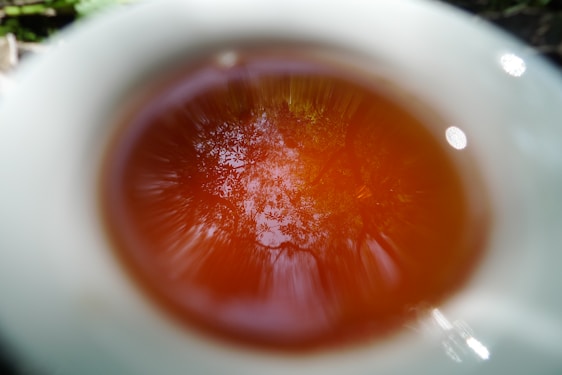 A reflection of trees and sky is visible on the surface of tea inside a white cup. The image captures a blurred, artistic effect where the outer edge of the cup is out of focus, drawing attention to the rich amber color of the tea and the intricate reflection within.