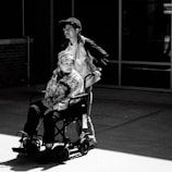 A black and white photo featuring a man standing behind a woman seated in a wheelchair. The woman is wearing a patterned jacket and a hat with text, while the man is wearing a jacket, shirt, and cap. They are in an outdoor setting with a brick wall and glass windows in the background.