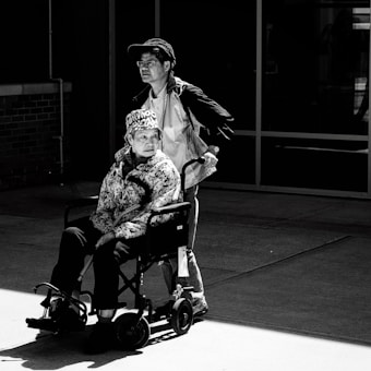 A black and white photo featuring a man standing behind a woman seated in a wheelchair. The woman is wearing a patterned jacket and a hat with text, while the man is wearing a jacket, shirt, and cap. They are in an outdoor setting with a brick wall and glass windows in the background.
