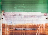 A weathered garage door with horizontal panels in varying shades of green, white, and rusty orange. Paint drips and smudges cover the surface, with visible textures and signs of decay. The metal appears aged, with areas of rust, adding an industrial, gritty character. A piece of wood is attached horizontally near the bottom.
