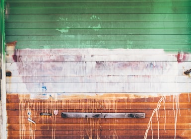 A weathered garage door with horizontal panels in varying shades of green, white, and rusty orange. Paint drips and smudges cover the surface, with visible textures and signs of decay. The metal appears aged, with areas of rust, adding an industrial, gritty character. A piece of wood is attached horizontally near the bottom.