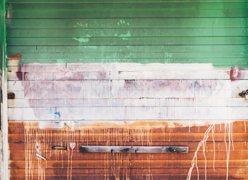 A weathered garage door with horizontal panels in varying shades of green, white, and rusty orange. Paint drips and smudges cover the surface, with visible textures and signs of decay. The metal appears aged, with areas of rust, adding an industrial, gritty character. A piece of wood is attached horizontally near the bottom.