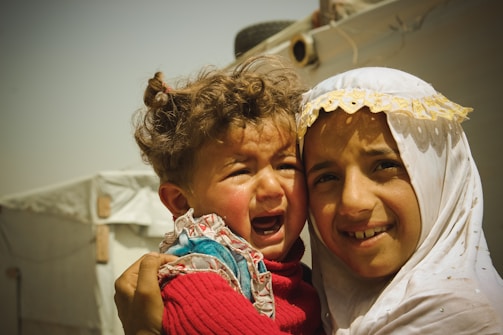 A young child with messy curly hair is crying while being held by an older child. The older child is smiling gently, wearing a white garment with yellow lace details. They are in an outdoor setting with a tent-like structure in the background.