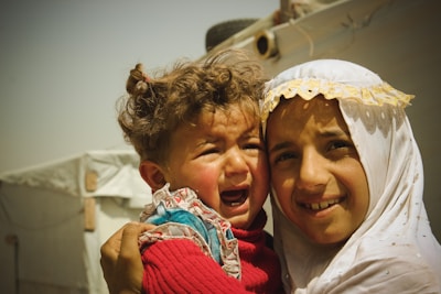 A young child with messy curly hair is crying while being held by an older child. The older child is smiling gently, wearing a white garment with yellow lace details. They are in an outdoor setting with a tent-like structure in the background.