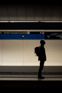 A silhouetted figure with a backpack stands on a subway platform near a sign reading 'Sortida Travessera Les Corts'. The platform is dimly lit with a contrasting bright wall behind. The architectural elements above suggest an underground station.