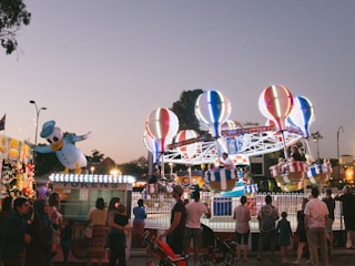 A lively carnival scene with a bright and colorful hot air balloon-themed ride in the center. Several people are seated in the ride's baskets, which are elevated off the ground. Surrounding the ride, there is a crowd of people, including families and children. A large inflatable character resembling a duck in a sailor hat is positioned to the left side. There are also lights and token stands visible, creating a festive atmosphere as the evening sky darkens.