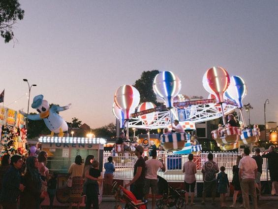 A lively carnival scene with a bright and colorful hot air balloon-themed ride in the center. Several people are seated in the ride's baskets, which are elevated off the ground. Surrounding the ride, there is a crowd of people, including families and children. A large inflatable character resembling a duck in a sailor hat is positioned to the left side. There are also lights and token stands visible, creating a festive atmosphere as the evening sky darkens.