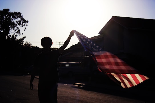 A silhouette of a person holding up an American flag in front of a bright sun. The flag is prominently displayed with its red and white stripes and blue field with stars. The background includes the outlines of houses and trees, creating a suburban setting.