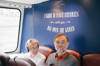 Two people are seated next to each other on a bus or a train. The background features a navy-blue wall with the words 'I want to make memories with you all over the world' written on it. One person is wearing headphones.