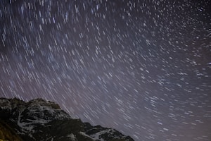 A night sky filled with star trails, creating curved lines across the image. The foreground features dark, rugged mountains partially covered in snow, adding contrast to the starry backdrop.