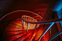 A spiral staircase with open metal steps and railings, arranged in a circular motion within a narrow, brick-walled space. The lighting creates a gradient effect with warm tones of red and orange blending with cooler blue hues, highlighting the texture of the brick and metal.