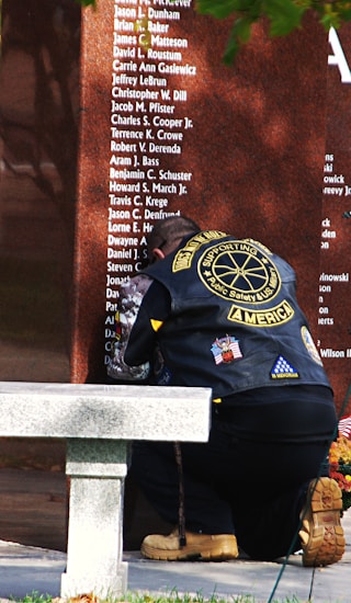 A person kneels in front of a reflective stone memorial wall, wearing a vest with various patches including American flags and 'SUPPORTING AMERICA' text. The person's head is bowed, indicating a moment of remembrance or respect. The wall has multiple names etched into its surface, suggesting it honors individuals. A stone bench and some greenery are visible in the foreground.