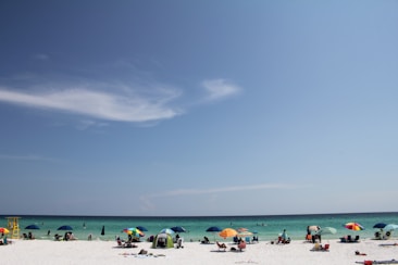A sunny beach scene with people relaxing under colorful umbrellas. The sandy shore is dotted with beachgoers, and the sea in the background is calm, transitioning from turquoise near the shore to deeper blue further out. A few clouds are scattered in the bright blue sky.