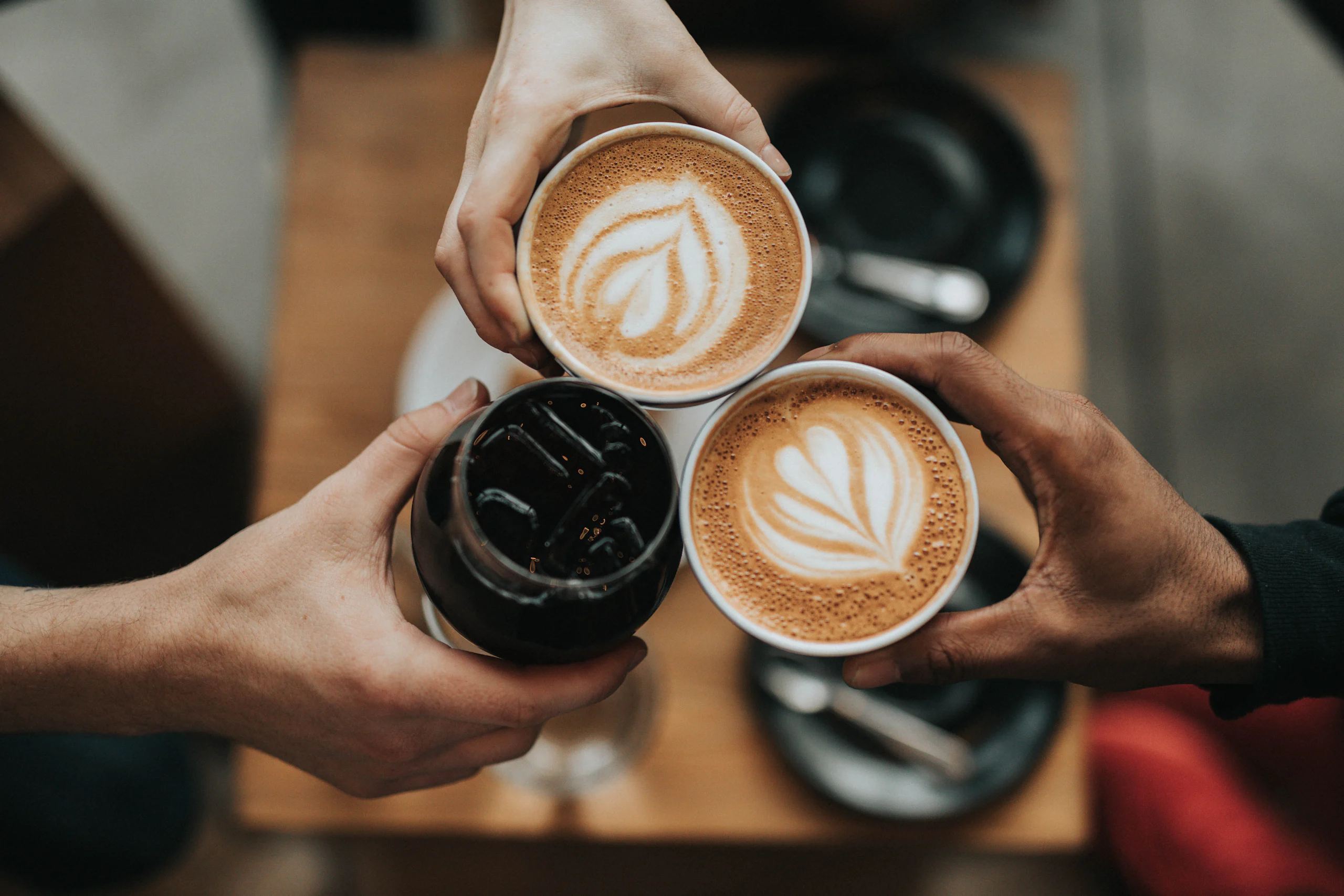 Trei mâini ciocnesc cești de cafea peste o masă de lemn