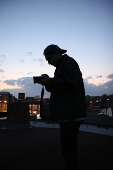 A silhouette of a person holding a camera stands against the backdrop of a twilight sky. The rooftop scene includes some cityscape elements and small urban structures. The sky is transitioning from day to night with a gradient of blue and hints of orange near the horizon.