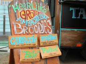 A wooden signboard with colorful and artistic text is displayed next to a food truck. The sign offers various types of sandwiches including Carpaccio, Martino, Brie, and Preparé. Each sandwich option lists its ingredients in smaller text below its name. The sign is adorned with lively, hand-painted letters.