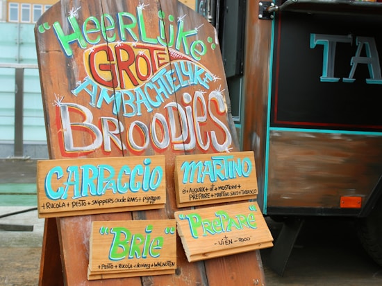 A wooden signboard with colorful and artistic text is displayed next to a food truck. The sign offers various types of sandwiches including Carpaccio, Martino, Brie, and Preparé. Each sandwich option lists its ingredients in smaller text below its name. The sign is adorned with lively, hand-painted letters.