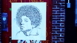 A piece of street art featuring a stylized portrait on a square canvas, mounted against a brick wall. The portrait comprises intricate patterns and text spiraling around the head, depicting a young person with voluminous hair. The surrounding environment includes a detailed red door and brick wall, with visible graffiti and urban elements.
