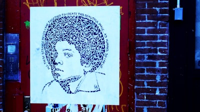 A piece of street art featuring a stylized portrait on a square canvas, mounted against a brick wall. The portrait comprises intricate patterns and text spiraling around the head, depicting a young person with voluminous hair. The surrounding environment includes a detailed red door and brick wall, with visible graffiti and urban elements.