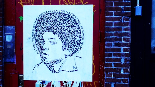 A piece of street art featuring a stylized portrait on a square canvas, mounted against a brick wall. The portrait comprises intricate patterns and text spiraling around the head, depicting a young person with voluminous hair. The surrounding environment includes a detailed red door and brick wall, with visible graffiti and urban elements.