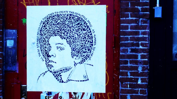 A piece of street art featuring a stylized portrait on a square canvas, mounted against a brick wall. The portrait comprises intricate patterns and text spiraling around the head, depicting a young person with voluminous hair. The surrounding environment includes a detailed red door and brick wall, with visible graffiti and urban elements.