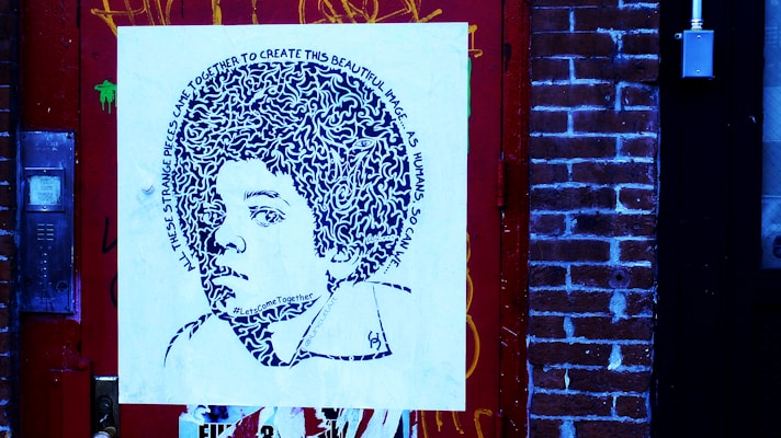 A piece of street art featuring a stylized portrait on a square canvas, mounted against a brick wall. The portrait comprises intricate patterns and text spiraling around the head, depicting a young person with voluminous hair. The surrounding environment includes a detailed red door and brick wall, with visible graffiti and urban elements.