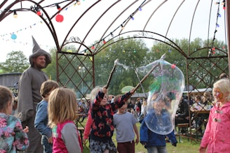 A group of children are playing with large soap bubbles outdoors, under a metal gazebo decorated with colorful string lights. A man dressed in a wizard-like costume stands nearby, watching the children. One child is using a stick to create the bubble, while others eagerly watch and try to catch it. The setting appears to be a festival or fair with other people and tents visible in the background. The scene is lively and playful, with a mix of vibrant colors and natural greenery.