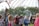 A group of children are playing with large soap bubbles outdoors, under a metal gazebo decorated with colorful string lights. A man dressed in a wizard-like costume stands nearby, watching the children. One child is using a stick to create the bubble, while others eagerly watch and try to catch it. The setting appears to be a festival or fair with other people and tents visible in the background. The scene is lively and playful, with a mix of vibrant colors and natural greenery.