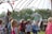 A group of children are playing with large soap bubbles outdoors, under a metal gazebo decorated with colorful string lights. A man dressed in a wizard-like costume stands nearby, watching the children. One child is using a stick to create the bubble, while others eagerly watch and try to catch it. The setting appears to be a festival or fair with other people and tents visible in the background. The scene is lively and playful, with a mix of vibrant colors and natural greenery.