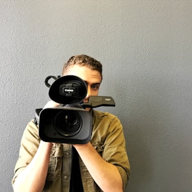 A person holds a professional video camera, pointing it directly at the viewer. The person has short, curly hair and wears a light brown jacket. The camera is positioned prominently in the center of the image, while the background is a plain, textured gray wall.