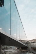 A modern architectural structure featuring a glass bridge connecting two concrete buildings. The bridge is elevated and surrounded by an urban setting, with reflections visible on the glass panels. The sky appears overcast, adding a muted tone to the environment.