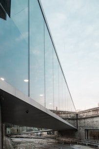 A modern architectural structure featuring a glass bridge connecting two concrete buildings. The bridge is elevated and surrounded by an urban setting, with reflections visible on the glass panels. The sky appears overcast, adding a muted tone to the environment.