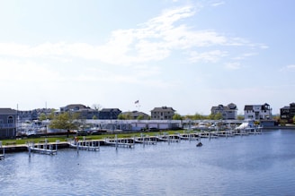 A serene waterfront scene featuring several modern houses along the shoreline, with a neatly lined row of docks extending into the calm water. The sky is mostly clear with a few scattered clouds. The area is verdant, with green lawns and trees visible near the water's edge. An American flag is visible flying near one of the houses.