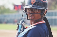 A person wearing a baseball helmet and jersey, holding a bat and looking towards the camera. The background includes a blurred view of a baseball field with a scoreboard.