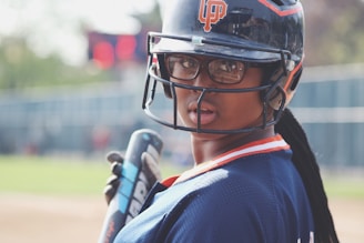 A person wearing a baseball helmet and jersey, holding a bat and looking towards the camera. The background includes a blurred view of a baseball field with a scoreboard.
